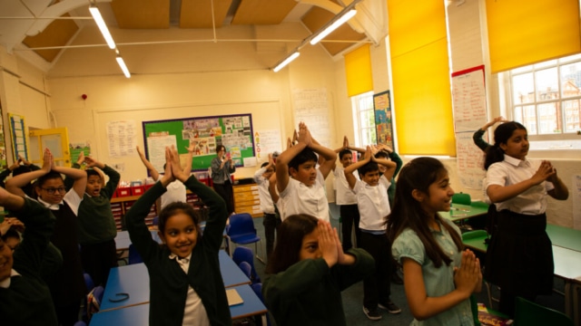 Photograph of children in a classroom with their hands together and up.