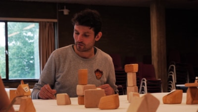 Photograph of a man drawing on a large piece of paper with wooden blocks in front of him.