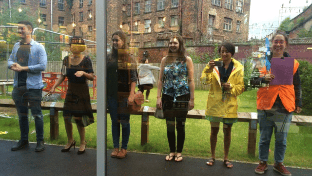 Photograph of the reflection of a group of people stood in a line in front of a glass window.