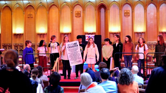 A group of young people are presenting on the red-carpeted stage of Guildhall's Great Hall. They are holding up a poster that reads: A poster that reads: 'The Future Charter. A group of 12-20 year-olds, friendly salute the City of London Corporation. And we declare that we want you to do everything that you can to create the best possible future for the 12-year-olds of today, and of the future. Keep us in mind as you make your mark on today. Your decisions today create the future of tomorrow.'